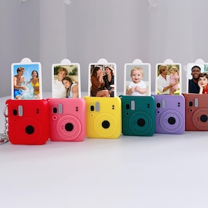 Puede incluir: Una colección de llaveros de cámara en miniatura de colores. Cada llavero presenta un color diferente: rojo, rosa, amarillo, verde, morado y marrón. Cada cámara contiene una pequeña foto. Los llaveros están dispuestos en fila sobre una superficie blanca.