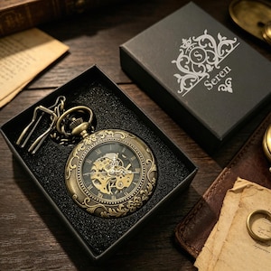 Peut inclure: Une montre de poche mécanique couleur bronze avec un boîtier orné de motifs floraux et un mécanisme à engrenages doré visible. La montre présente des chiffres romains sur un cadran noir et est présentée dans une boîte noire marquée Seren.
