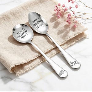 Op de afbeelding: Twee zilverkleurige metalen lepels met gegraveerde tekst en afbeeldingen. Eén lepel bevat de tekst Aaron's ice cream spoon en een ijshoorntje. De andere lepel bevat de tekst Jane's Wedding Shower en een vogel met een hartje als icoon.