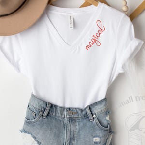 May include: White V-neck t-shirt with the word "magical" in red script. The shirt is paired with denim shorts. A tan hat hangs in the background. The shirt is a casual, everyday wear item.