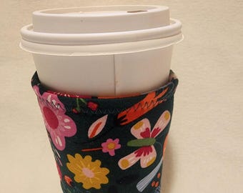 May include: A white coffee cup with a green, pink, yellow, and orange floral print fabric sleeve. The sleeve has a stitched seam around the top and bottom.