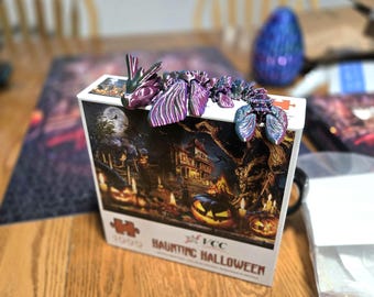 Może przedstawiać: Puzzle "Haunting Halloween" z 1000 elementami, z upiornym obrazem nawiedzonego domu, dyni i pełni księżyca. Ozdobny, opalizujący fioletowo-zielony smok siedzi na pudełku z puzzlami.