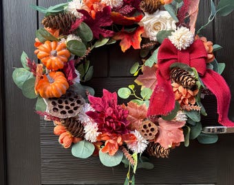 May include: A fall-themed wreath with a variety of decorative elements. It features orange pumpkins, pinecones, and faux flowers in shades of red, orange, and white. A red burlap bow adds a festive touch. The wreath is displayed on a dark wooden door.