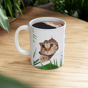 Puede incluir: Taza de café de cerámica blanca con un gato atigrado marrón asomándose por un agujero en el lateral de la taza. El gato tiene ojos verdes y está rodeado de hojas verdes.