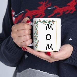 Puede incluir: Taza de cerámica blanca con un diseño floral y la palabra "MOM" en letras negras dentro de un marco dorado.