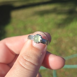 May include: A silver ring with a light green, oval-shaped gemstone. The ring has a detailed band with three horizontal lines and a scalloped setting around the stone. The ring is held up by a person's fingers.