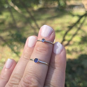 May include: Two silver rings with small, round, blue gemstones are displayed on a hand. The rings have a simple, delicate design, with the gemstones set in a minimalist style. The background is blurred, with green foliage.