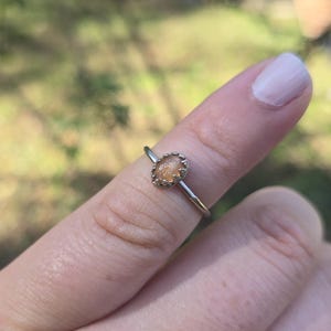 May include: A silver ring with an oval, light-colored gemstone. The ring is on a finger, with a blurred green background. The gemstone is set in a decorative bezel.