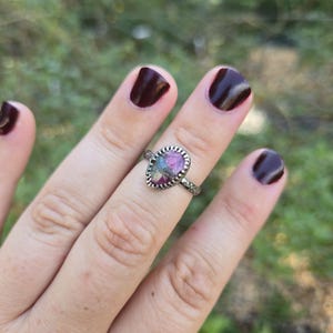 May include: A silver ring featuring an oval-shaped, multi-colored gemstone with shades of pink, green, and grey. The stone is set in a serrated metal bezel, and the ring band has a textured, patterned surface along its outer circumference.
