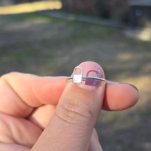 May include: A silver ring featuring a padlock design. The ring has a thin band and a small, rectangular padlock charm. The ring is held up by a finger, with a blurred background of green and brown.