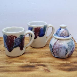 May include: A ceramic mug and sugar bowl set. The mugs and sugar bowl are off-white with a glaze of red, blue, and green. The sugar bowl has a lid. The set is on a wooden surface.