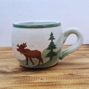 May include: A ceramic mug with a white base, featuring a brown moose silhouette and a green pine tree design. The rim and handle have a green speckled finish. The mug is designed for hot beverages.
