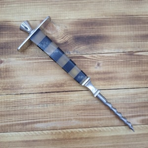 May include: A silver-toned corkscrew with a black and tan checkered handle. The corkscrew has a flat top and a twisted, pointed end. The handle is rectangular and tapers towards the bottom. The item is displayed on a wooden surface.