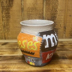 May include: A ceramic mug with a patchwork design featuring the logos of popular candy brands, including Reese's, M&M's, and Kit Kat.