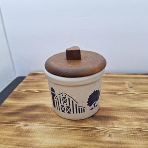 May include: A cream-colored ceramic jar with a wooden lid. The jar features a blue barn and tree design. The lid has a small, square wooden handle. The jar is sitting on a wooden surface.