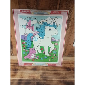 May include: A framed Playskool My Little Pony wooden board puzzle. The puzzle features a white pony with blue hair and a pink bow, a pink castle, and a purple creature. The puzzle is for ages 2 to 5 years.