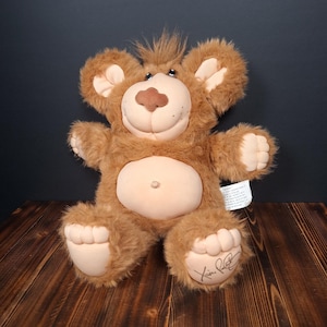 May include: A brown plush teddy bear with a soft, light-colored fabric face, belly, ears, and paws. The bear features a tuft of brown hair on its head, a dark brown nose, and a signature on the bottom of its left foot. A white tag is attached to the side.