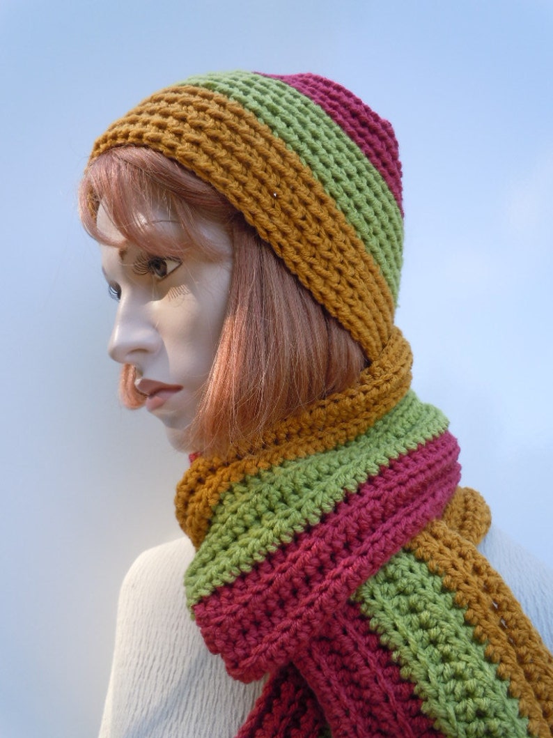 Op de afbeelding: Een gebreide set van muts en sjaal in tinten mosterdgeel, groen en bordeauxrood. De muts is een slouchy beanie-stijl en de sjaal is lang en breed.