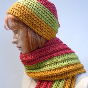 Op de afbeelding: Een gebreide set van muts en sjaal in tinten rood, groen en oranje. De muts is een slouchy beanie-stijl en de sjaal is lang en gestreept.