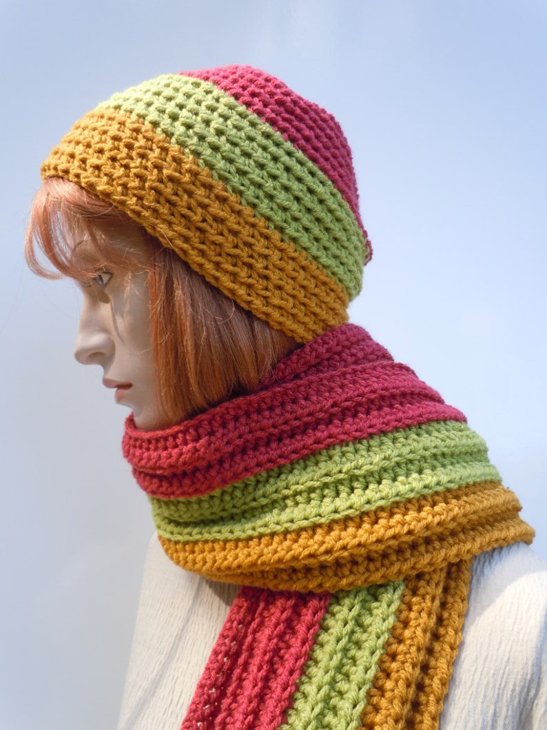 Op de afbeelding: Een gebreide set van muts en sjaal in tinten rood, groen en oranje. De muts is een slouchy beanie-stijl en de sjaal is lang en gestreept.