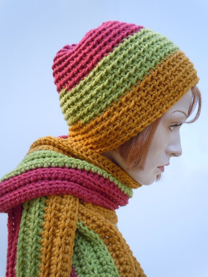 Op de afbeelding: Een gehaakte set van muts en sjaal in tinten groen, oranje en roze. De muts is slouchy en de sjaal is lang en breed.