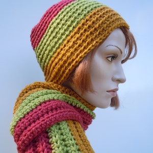Op de afbeelding: Een gebreide set van muts en sjaal in tinten rood, groen en oranje. De muts is een slouchy beanie-stijl en de sjaal is lang en breed.