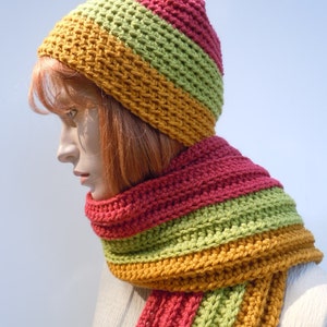 Op de afbeelding: Een gebreide set van muts en sjaal in tinten groen, oranje en rood. De muts is een slouchy beanie-stijl en de sjaal is lang en breed.