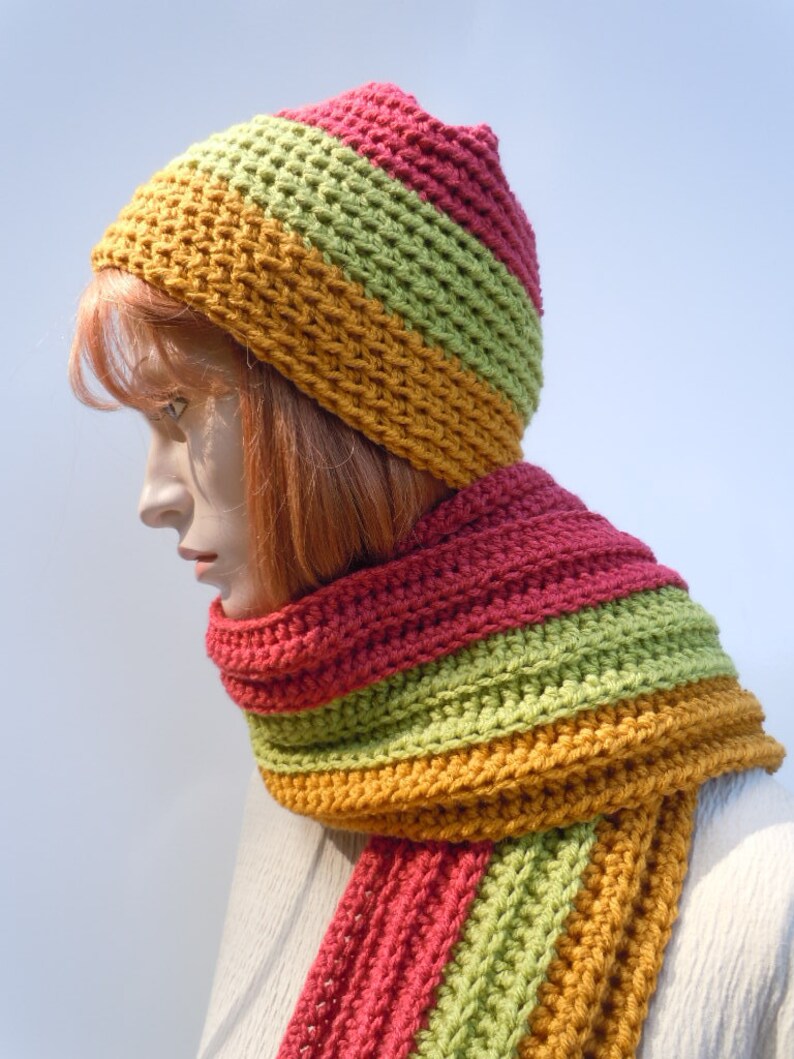 Op de afbeelding: Een gebreide set van muts en sjaal in tinten groen, oranje en rood. De muts is een slouchy beanie-stijl en de sjaal is lang en breed.