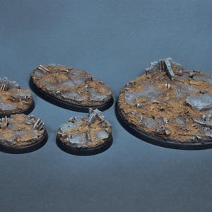 May include: Four different sizes of oval-shaped miniature bases for tabletop games. The bases are made of a brown resin and feature a textured surface with gray and brown details. The bases are designed to represent a ruined cityscape.