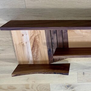May include: A wall-mounted shelf unit made of walnut and maple wood. The shelf has a geometric design with a curved lower shelf and multiple hooks on the right side.