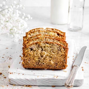 Könnte beinhalten: Nahaufnahme eines weißen Schneidebretts mit vier Scheiben Bananenbrot darauf. Das Brot ist goldbraun und hat eine leicht knusprige Kruste. Ein silbernes Buttermesser liegt rechts neben dem Brot auf dem Brett. Das Brett liegt auf einer weißen Marmorfläche.
