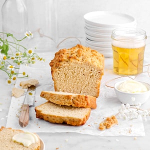 Puede incluir: Un pan dorado parcialmente rebanado, con dos rebanadas delante. Un plato pequeño con una rebanada de pan y mantequilla, un tarro de cerveza y un cuenco de mantequilla también están presentes. La escena se desarrolla sobre una superficie blanca con flores.