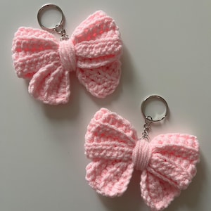 May include: Two handmade pink crochet bow keychains. Each bow is attached to a silver-colored keyring. The bows are made of soft, textured yarn, and the design is a classic bow shape.