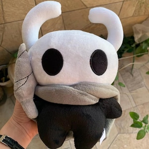 Può includere: Peluche bianco e nero di un personaggio con occhi grandi e un mantello. Il giocattolo è fatto di tessuto morbido e coccoloso ed è alto circa 25 cm.