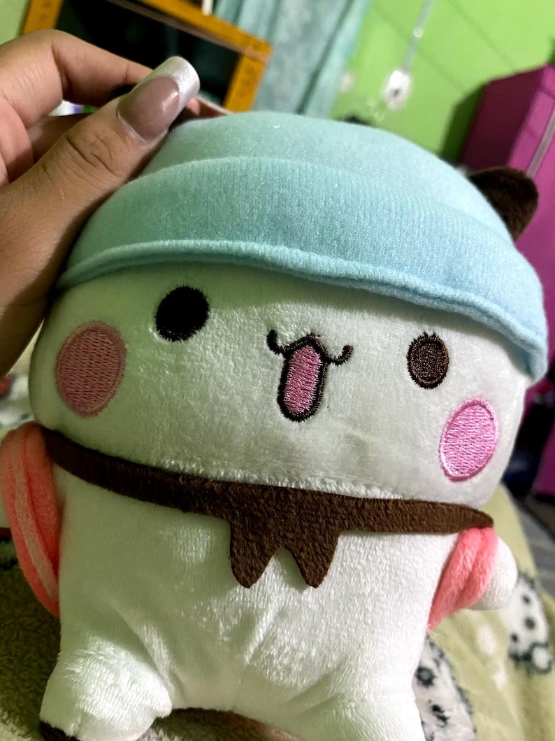 Puede incluir: Juguete de peluche blanco con un sombrero azul, ojos marrones, mejillas rosadas y un bigote marr&oacute;n. El juguete lleva una bufanda marr&oacute;n y tiene detalles rosas y blancos.