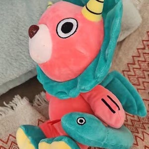 Op de afbeelding: Een pluche speelgoed in de vorm van een roze en turquoise wezen met een gele hoorn en een vis in zijn mond. Het speelgoed zit op een rood-wit gepatroneerd oppervlak.