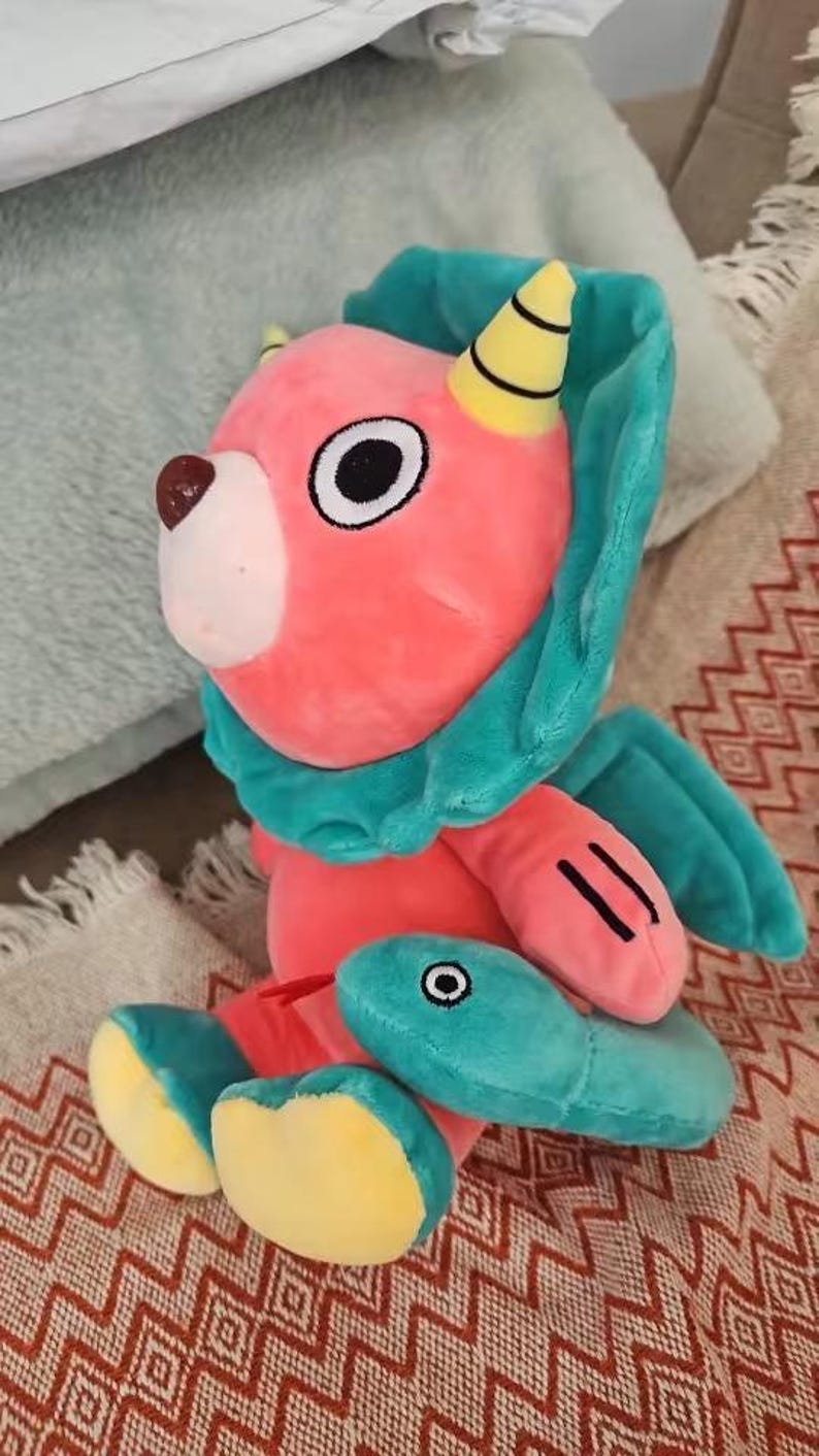 Op de afbeelding: Een pluche speelgoed in de vorm van een roze en turquoise wezen met een gele hoorn en een vis in zijn mond. Het speelgoed zit op een rood-wit gepatroneerd oppervlak.