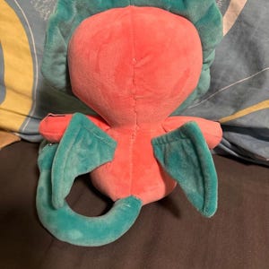 Op de afbeelding: Een roze pluche speelgoed met turquoise vleugels en een staart. Het speelgoed heeft een gestikte naad op de rug en een rond hoofd.