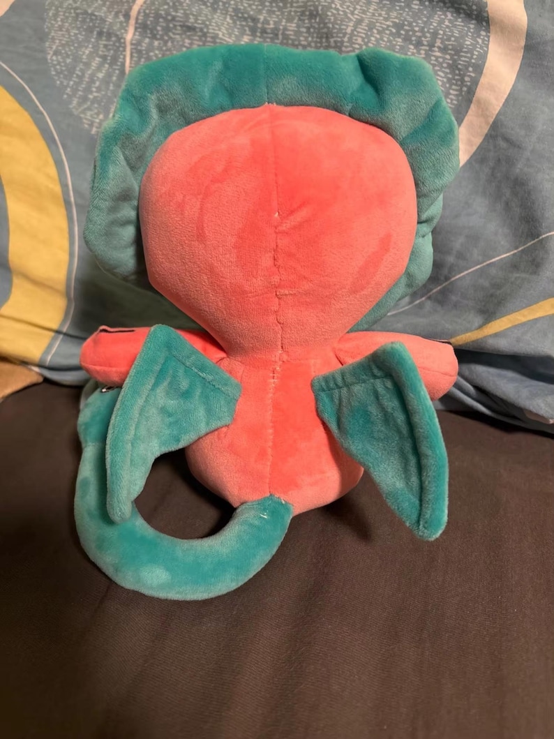 Op de afbeelding: Een roze pluche speelgoed met turquoise vleugels en een staart. Het speelgoed heeft een gestikte naad op de rug en een rond hoofd.