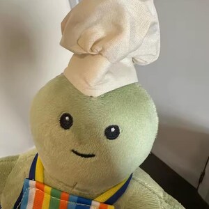 Puede incluir: Un peluche verde con un gorro de chef y un delantal a rayas de colores. El juguete tiene ojos negros de bot&oacute;n y una sonrisa cosida. El delantal presenta rayas verticales en varios colores.
