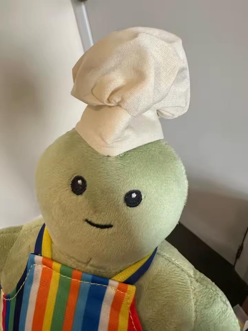 Puede incluir: Un peluche verde con un gorro de chef y un delantal a rayas de colores. El juguete tiene ojos negros de bot&oacute;n y una sonrisa cosida. El delantal presenta rayas verticales en varios colores.