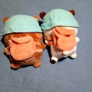 Puede incluir: Dos juguetes de peluche con sombreros azul claro y caras marrones. Cada juguete tiene una peque&ntilde;a mochila naranja.