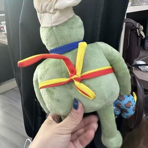 Puede incluir: Un peluche verde con forma de rana, con un gorro de chef blanco, un cuello azul y una faja roja y amarilla. La rana est&aacute; sujeta por una persona. Un peque&ntilde;o juguete azul y estampado es visible en el fondo.