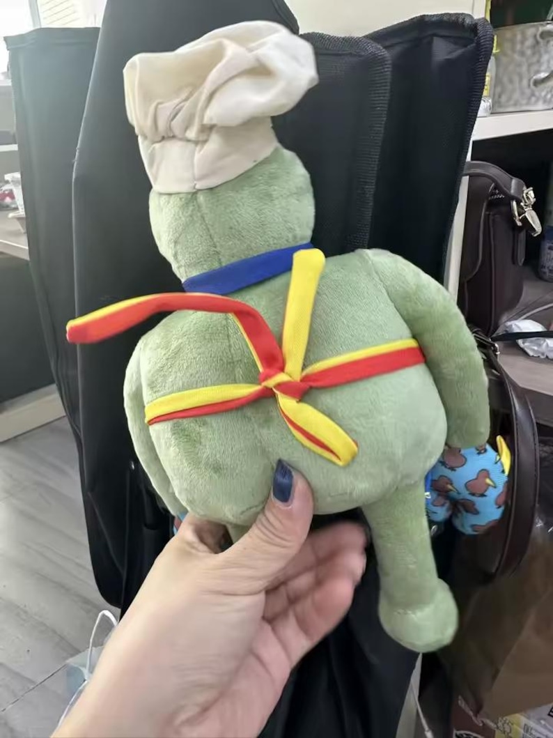 Puede incluir: Un peluche verde con forma de rana, con un gorro de chef blanco, un cuello azul y una faja roja y amarilla. La rana est&aacute; sujeta por una persona. Un peque&ntilde;o juguete azul y estampado es visible en el fondo.
