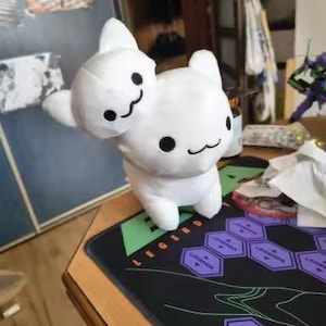 Puede incluir: Un peluche blanco con dos cabezas, cada una con ojos negros y una boca sonriente. El juguete est&aacute; sobre una alfombrilla de rat&oacute;n para juegos negra y verde con dise&ntilde;os hexagonales morados y la palabra "LEGEND" en naranja.