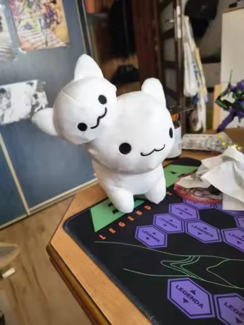 Puede incluir: Un peluche blanco con dos cabezas, cada una con ojos negros y una boca sonriente. El juguete est&aacute; sobre una alfombrilla de rat&oacute;n para juegos negra y verde con dise&ntilde;os hexagonales morados y la palabra "LEGEND" en naranja.