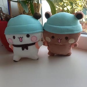 Puede incluir: Dos peluches con gorros azules. El peluche de la izquierda es blanco con una pajarita marr&oacute;n y mejillas rosas. El peluche de la derecha es marr&oacute;n con orejas marrones y mejillas rosas.
