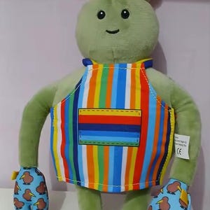 Puede incluir: Un personaje de peluche verde con un gorro de chef y un delantal a rayas de colores. El personaje tiene guantes con estampado de kiwi y una cara sonriente. Un juguete de peluche divertido y caprichoso.