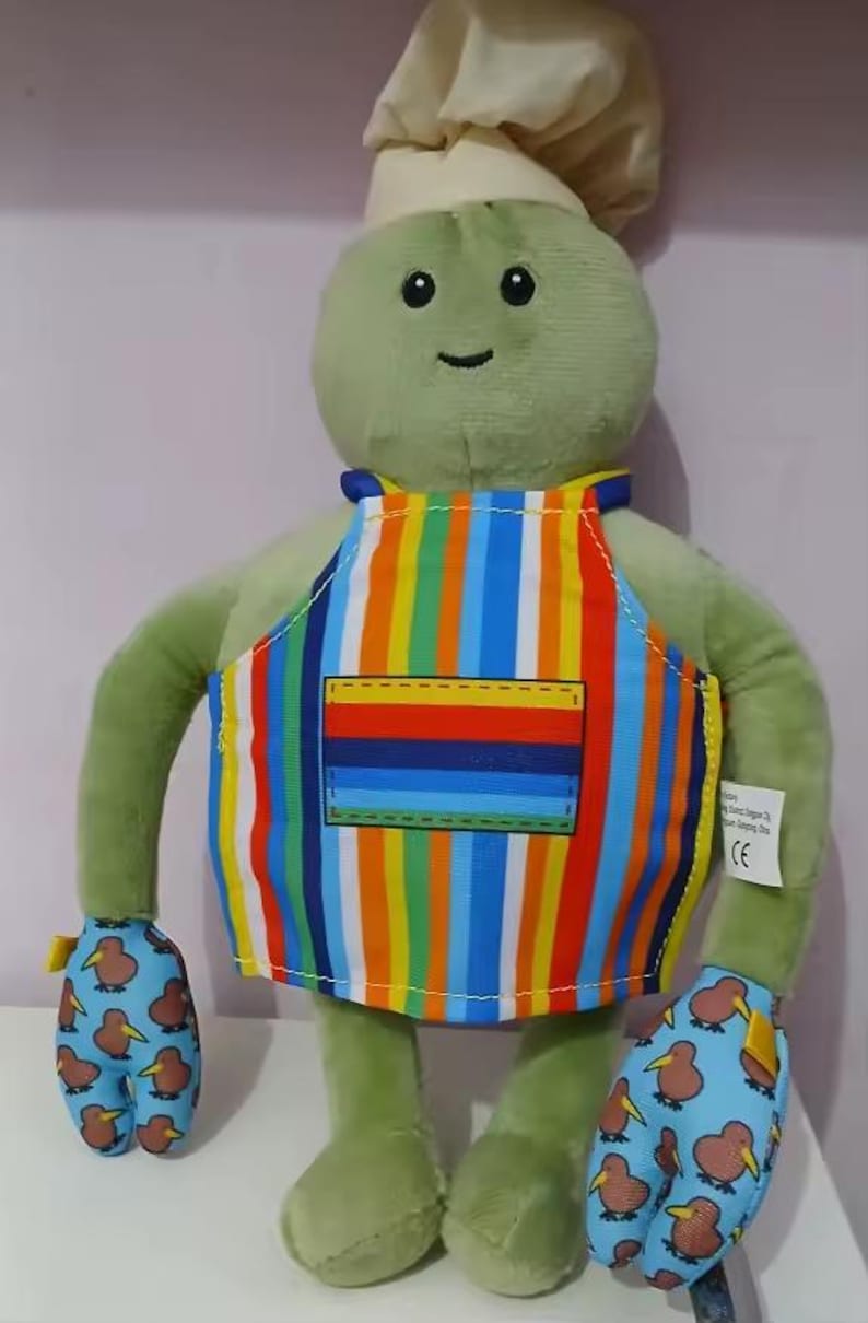 Puede incluir: Un personaje de peluche verde con un gorro de chef y un delantal a rayas de colores. El personaje tiene guantes con estampado de kiwi y una cara sonriente. Un juguete de peluche divertido y caprichoso.