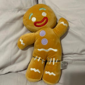 Puede incluir: Un juguete de peluche de hombre de jengibre con una cara sonriente, ojos blancos, boca roja y botones morados. El juguete está hecho de tela suave y marrón y tiene un diseño cosido.